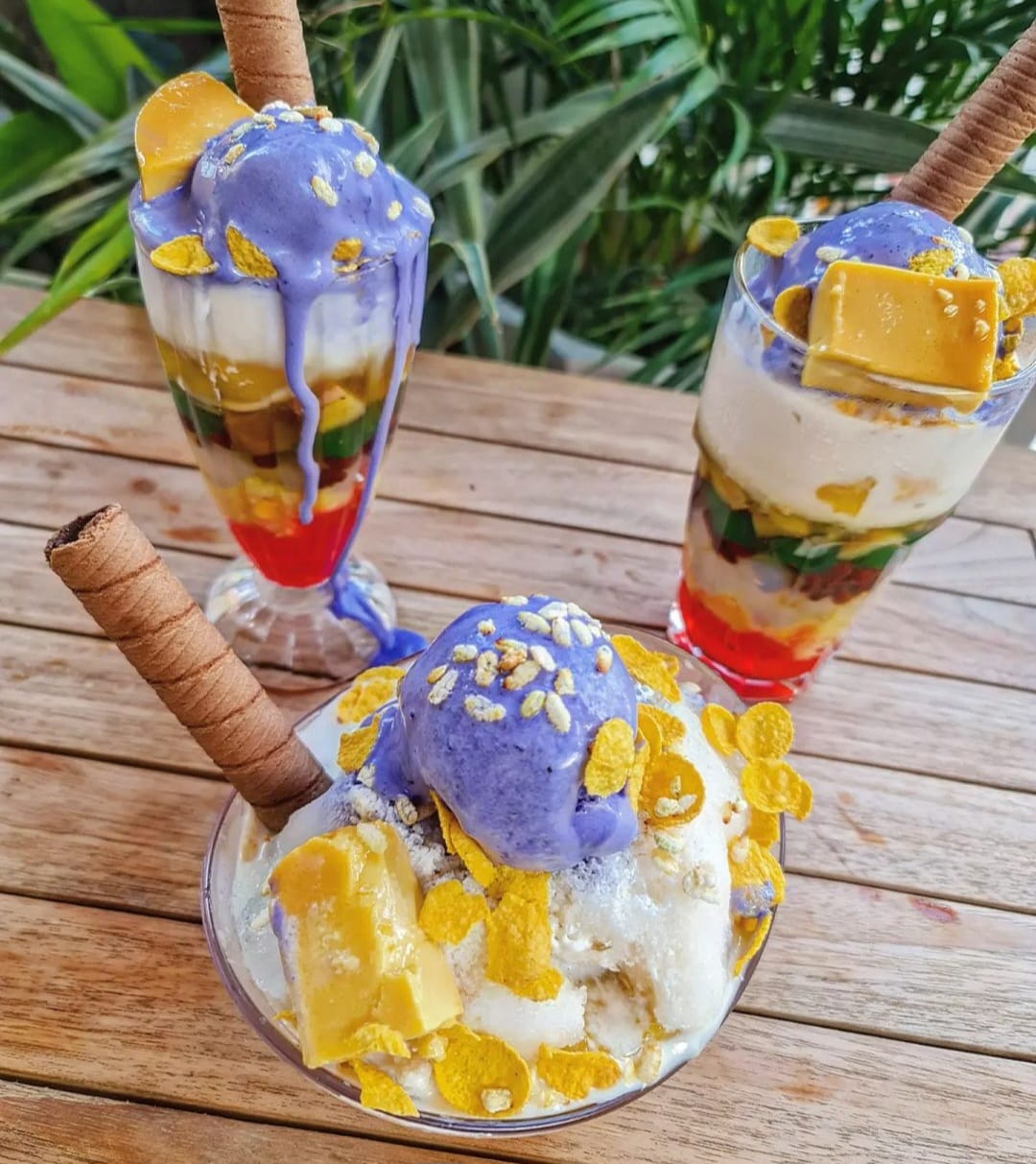 Two glasses of halo halo topped with melting ube ice cream, leche flan, cornflakes, and wafer rolls, served outdoors on a wooden table with tropical greenery in the background. 